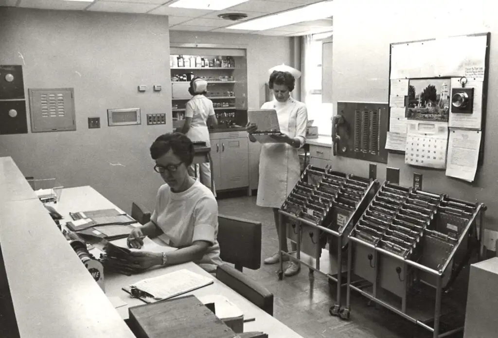 Black-and-white photo of a hospital nurses’ station from decades past, showing three nurses working with paper charts and medical files in a clinical office setting.